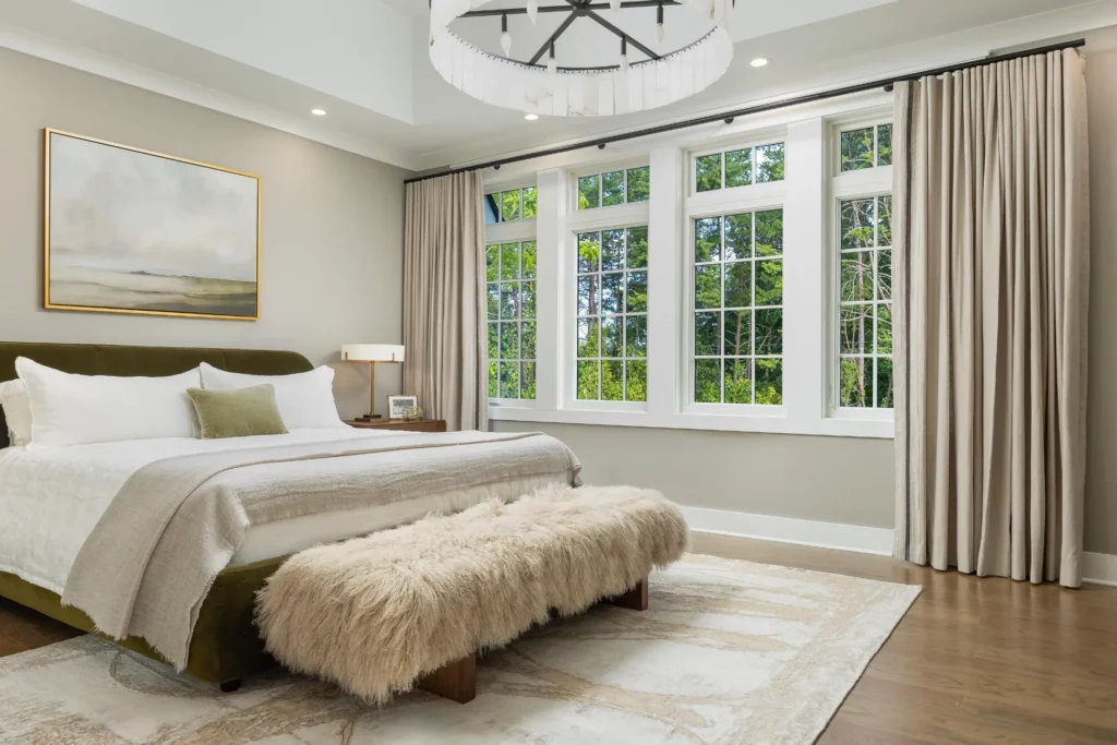 A bedroom designed by an interior designer in Chattanooga features a large bed with white bedding, a green bed frame, a fur bench, and modern chandelier. Natural light pours in through large windows adorned with beige curtains, highlighting the artwork on the wall.