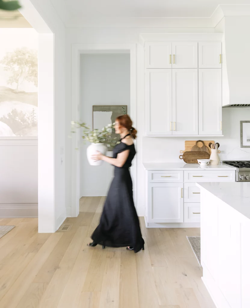 A person in a black dress walks through a bright, modern kitchen with white cabinets and hardwood floors, carrying a white pot with green foliage.