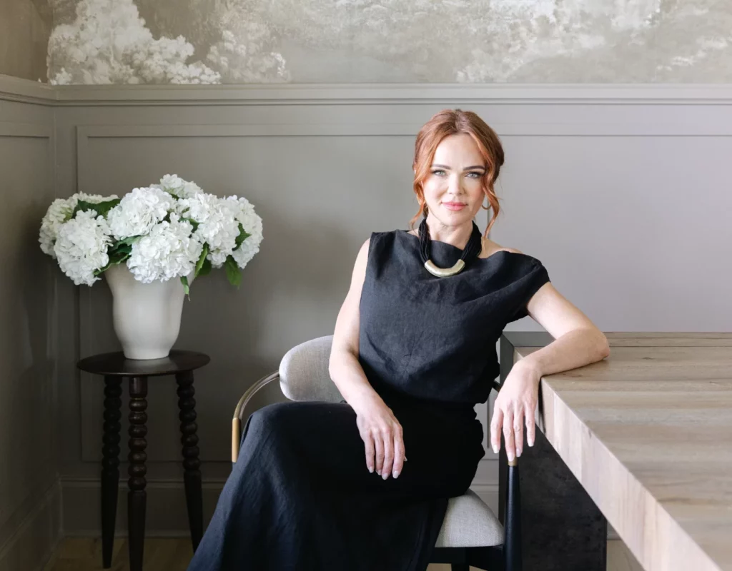 A woman in a black dress and earrings sits on a chair next to a wooden table. There's a vase with white flowers on a small table nearby. The wall behind has a landscape mural.