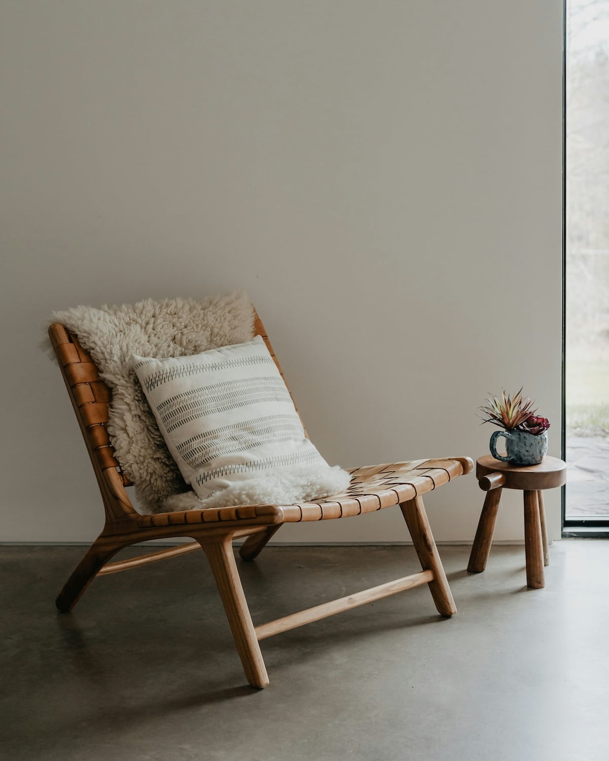 A wooden chair with a cushion and a fringed throw sits beside a small wooden table holding a potted plant in a minimalist room.