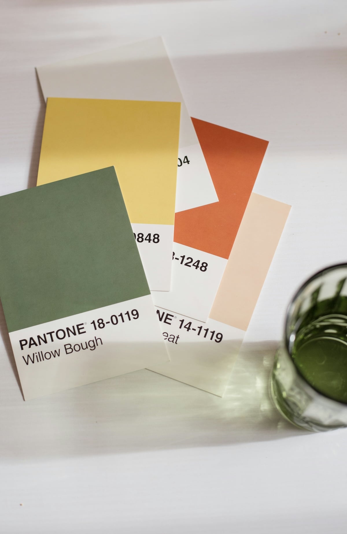 A selection of Pantone color swatches, including Willow Bough, Brushed Nickle, and Oak Buff, displayed on a white surface next to a green glass.