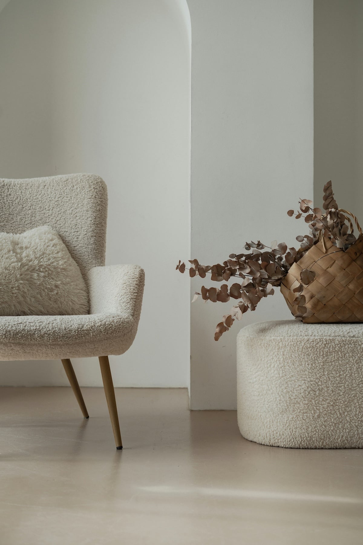 A cozy beige armchair with a matching pillow sits beside a textured ottoman and a woven basket containing dried leaves, against a minimalist cream-colored wall with an archway.