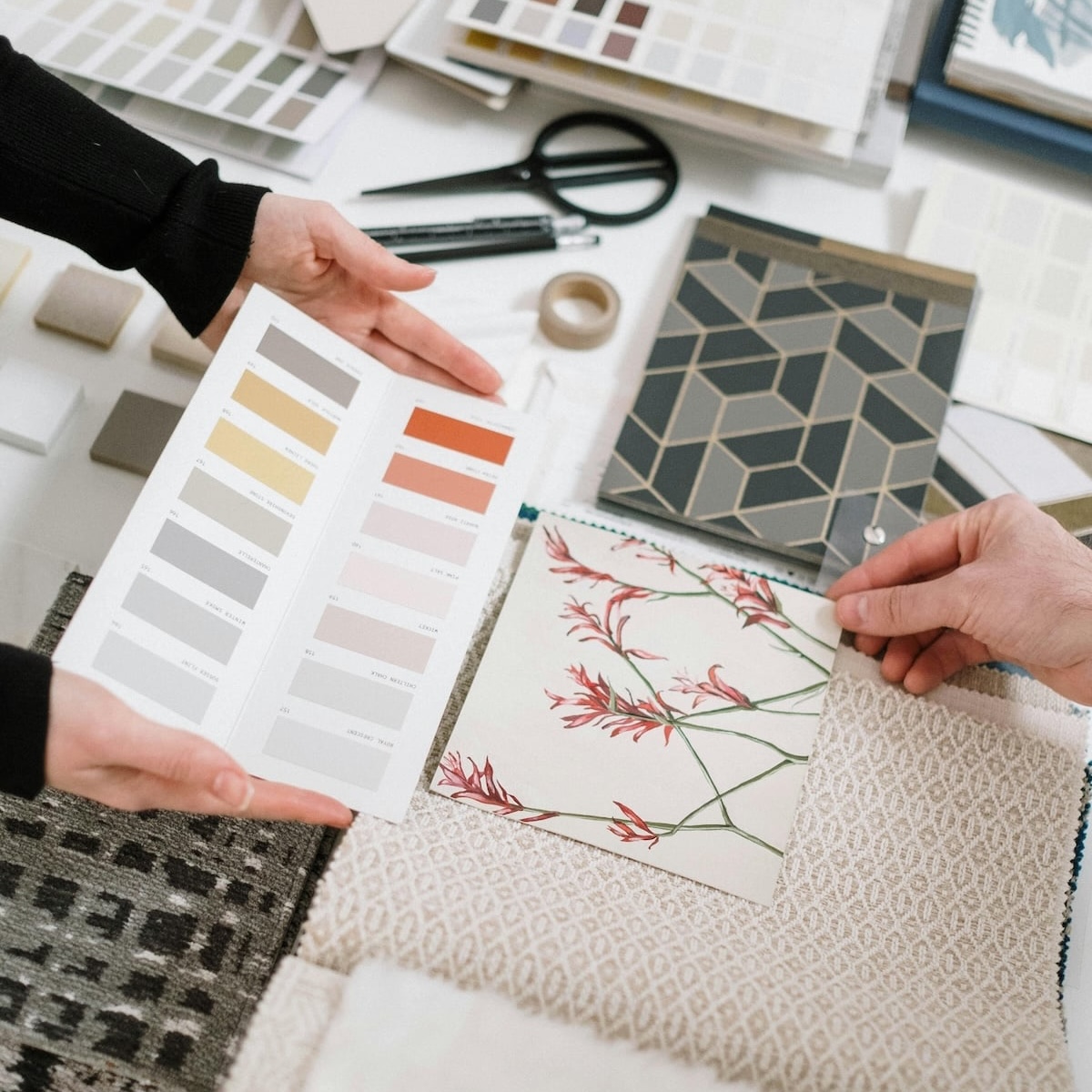 Two people are selecting materials, including color swatches, fabric samples, and tile designs, from an assortment spread on a table.