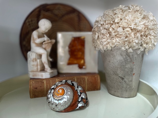 A white statue of a seated figure, a square glass item, a flowerpot, a decorative shell, and a closed book are arranged on a tabletop.