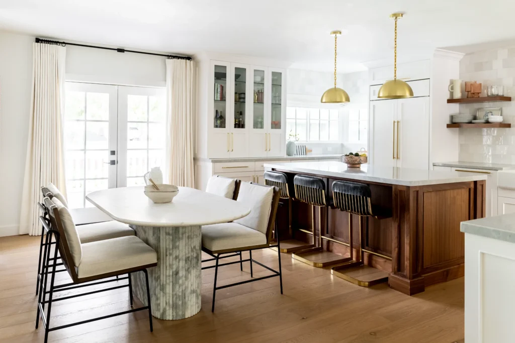 A modern kitchen with a marble island, three gold pendant lights, black and gold barstools, a wooden dining table with white chairs, light hardwood floors, white cabinets, and glass doors to the outside.